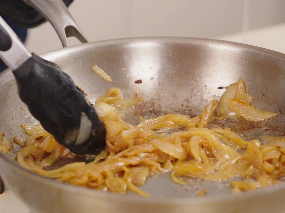 Tongs stirring a thick, jammy pile of deep golden caramelized onions in a shimmering stainless steel pan.
