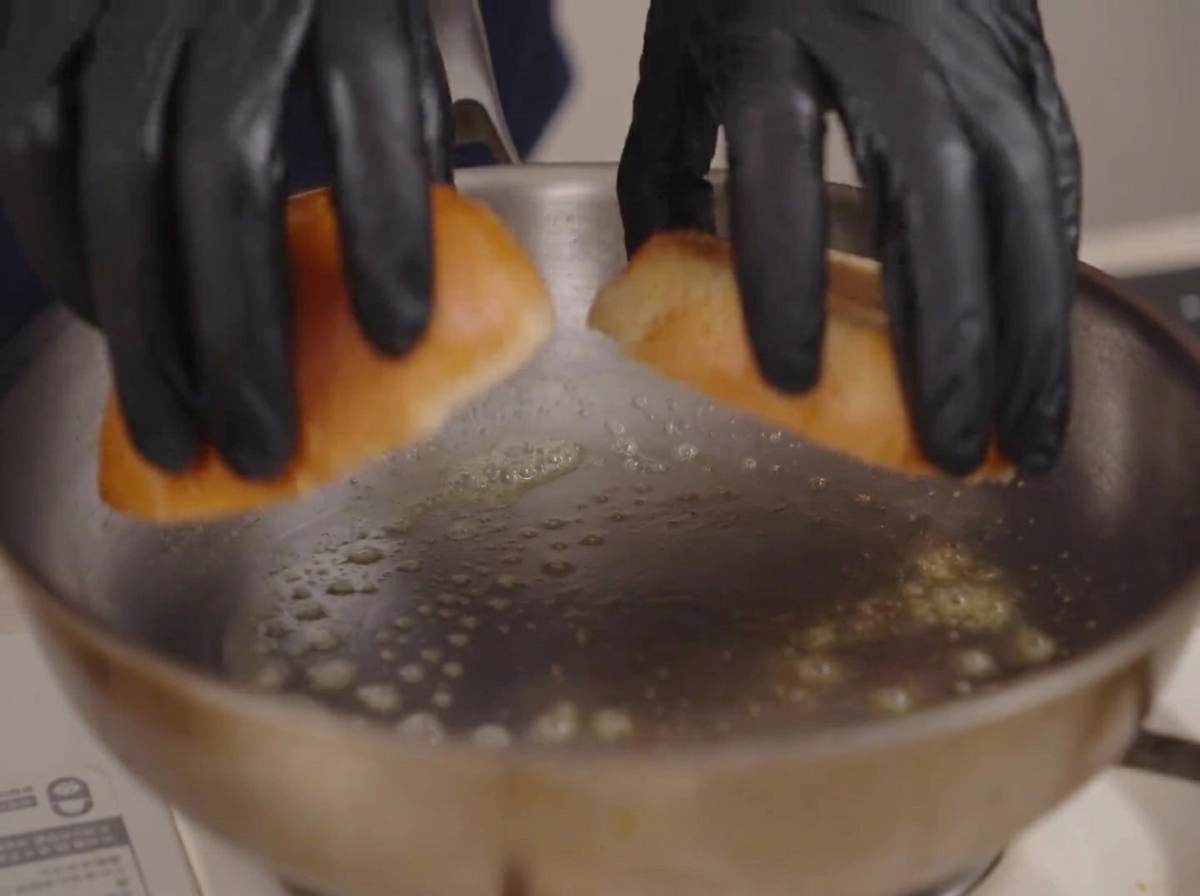 A pair of gloved hands placing two brioche bun halves into a stainless steel pan with bubbling melted butter.