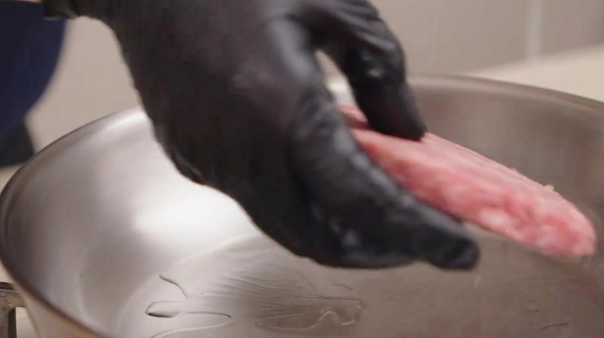 A hand wearing a black kitchen glove carefully placing a raw, marbled beef patty into a shimmering stainless steel skillet with oil.