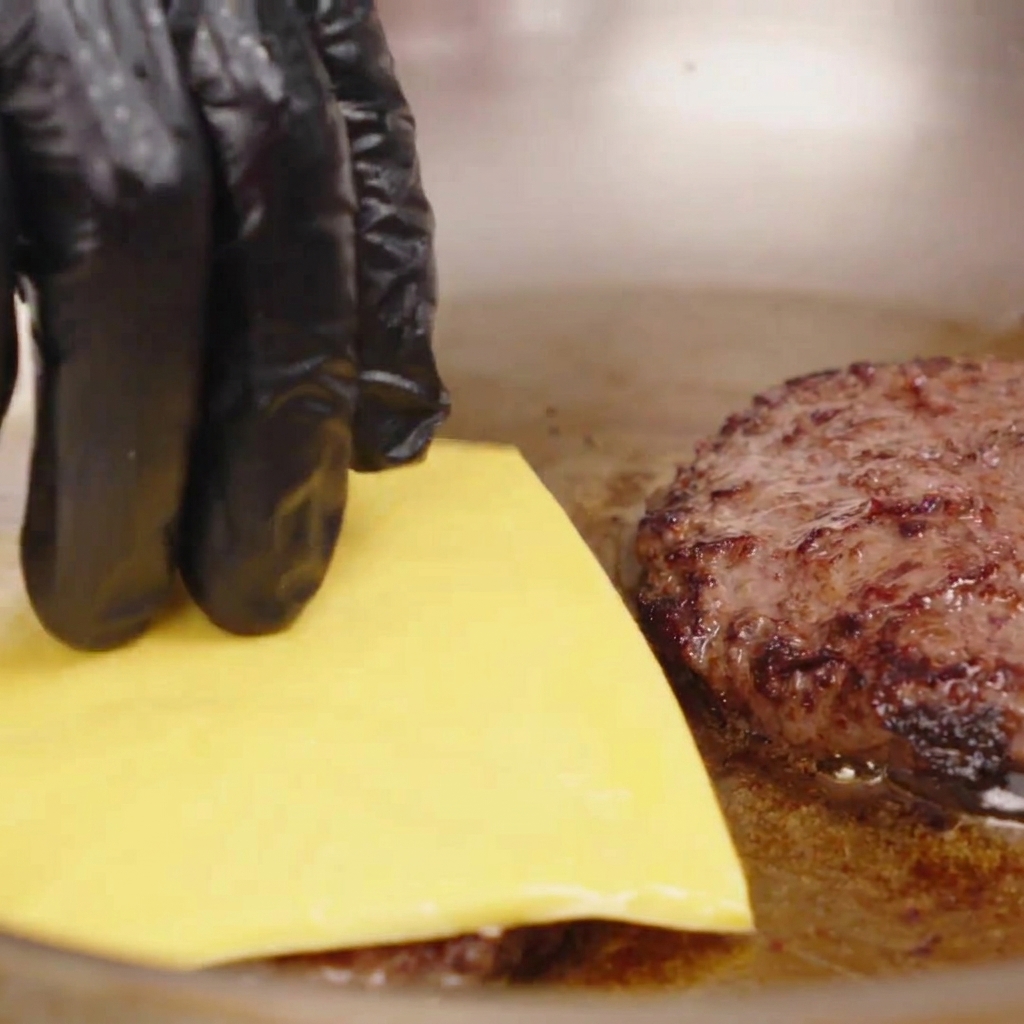 A gloved hand placing a square slice of yellow cheese onto a hot, seared beef patty in a pan.