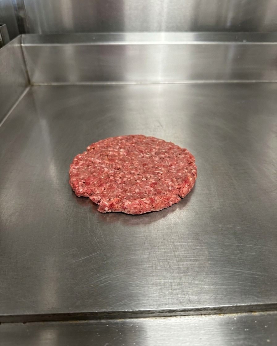 A flattened raw beef patty on a metal surface, seasoned with visible grains of salt and pepper.