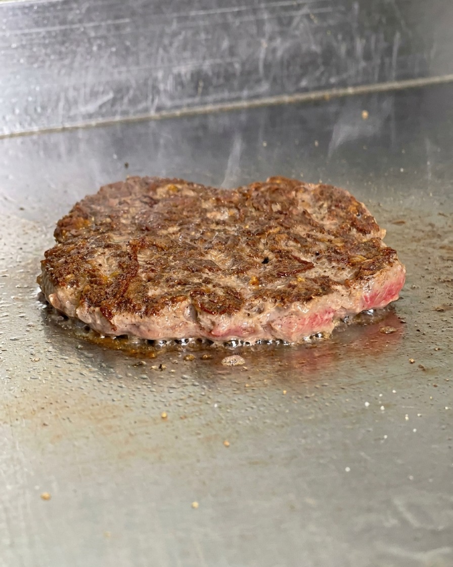A cooked burger patty flipped over on a griddle showing a dark brown, crispy seared crust with fat bubbling around the edges.