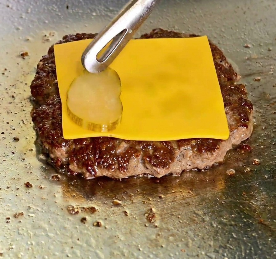 A slice of melted yellow cheddar cheese on a beef patty with tongs placing a pickle slice on top.