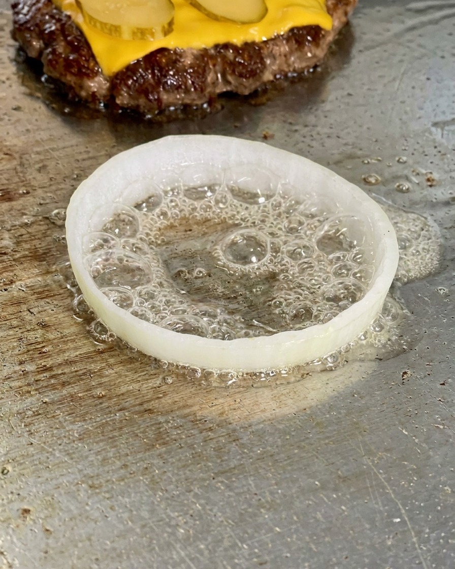 A slice of white onion on a griddle with a raw egg frying inside it, surrounded by bubbling oil.