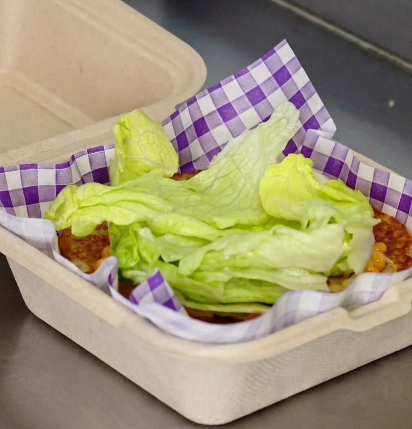 Fresh green lettuce leaves being stacked on top of a toasted bun with red sauce inside a purple checkered paper liner.