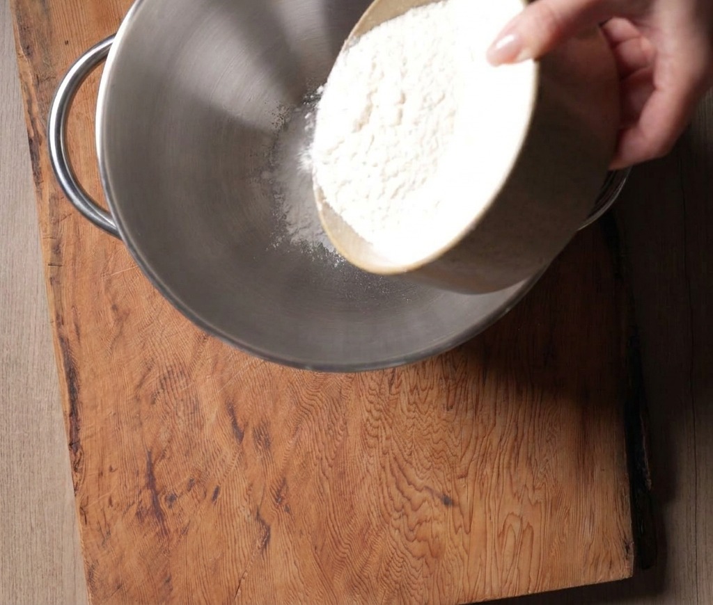 Fine white high-gluten flour being poured from a ceramic bowl into a large stainless steel stand mixer bowl.