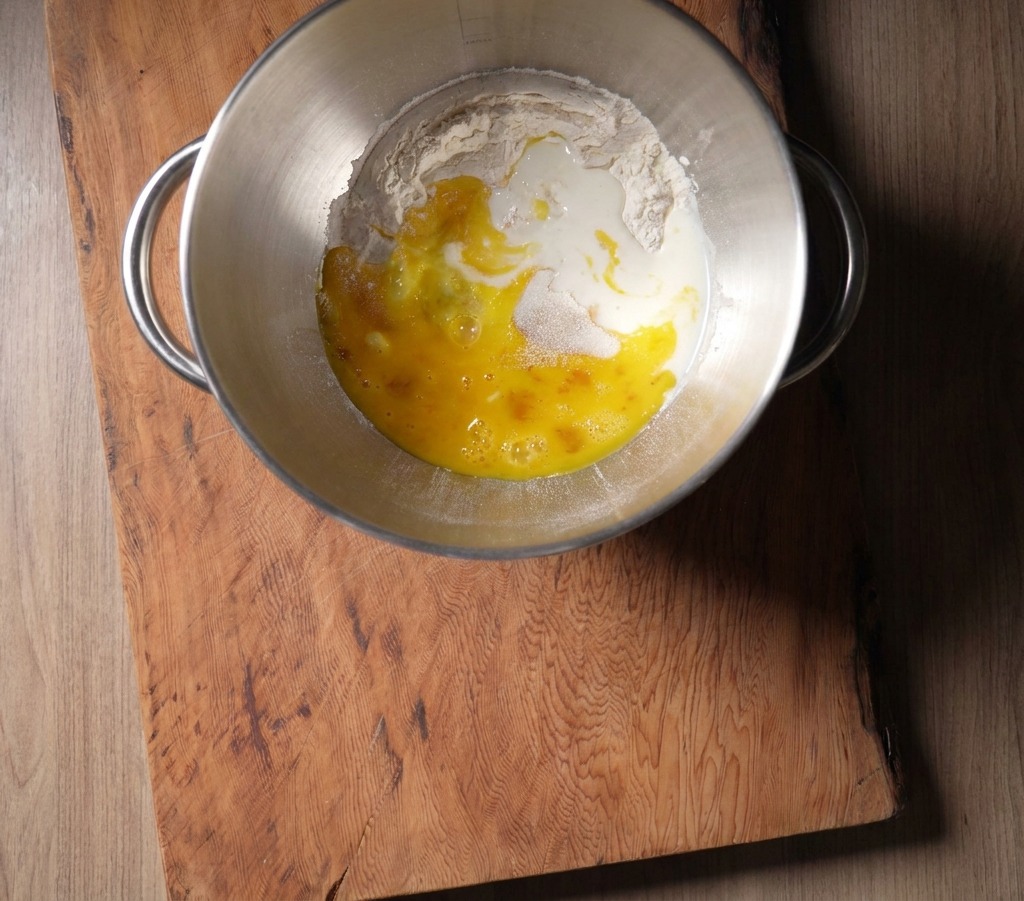 A stainless steel mixing bowl containing flour and a bright yellow egg mixture, with fresh milk being poured in from above.