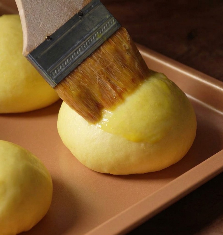 A close-up shot of a wooden pastry brush applying a golden yellow egg wash onto a smooth, rounded brioche dough ball on a copper-colored baking sheet.