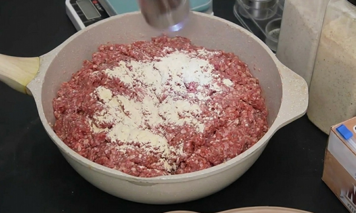 An electric stainless steel spice mill grinding black pepper over seasoned raw meat in a pan.