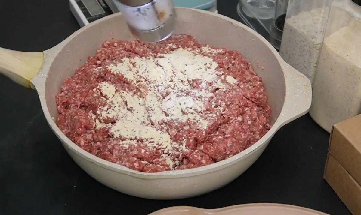 A person in a black glove using an electric grinder to add sea salt to raw ground beef.