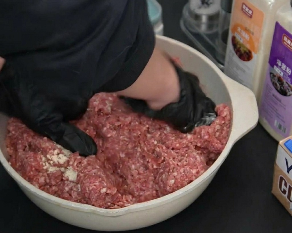 Hands wearing black gloves mixing and kneading seasoned raw ground beef in a light-colored pan.