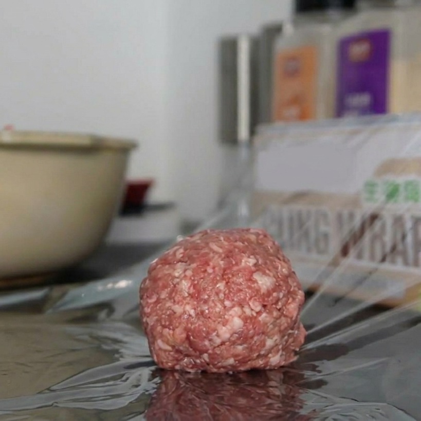 A single, round portion of seasoned ground beef resting on a piece of clear cling wrap on a countertop.
