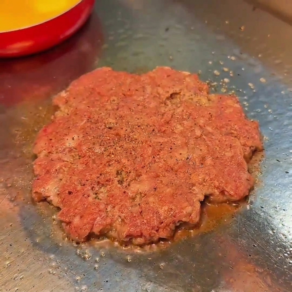 A raw beef patty cooking on a flat grill, seasoned with salt and black pepper.