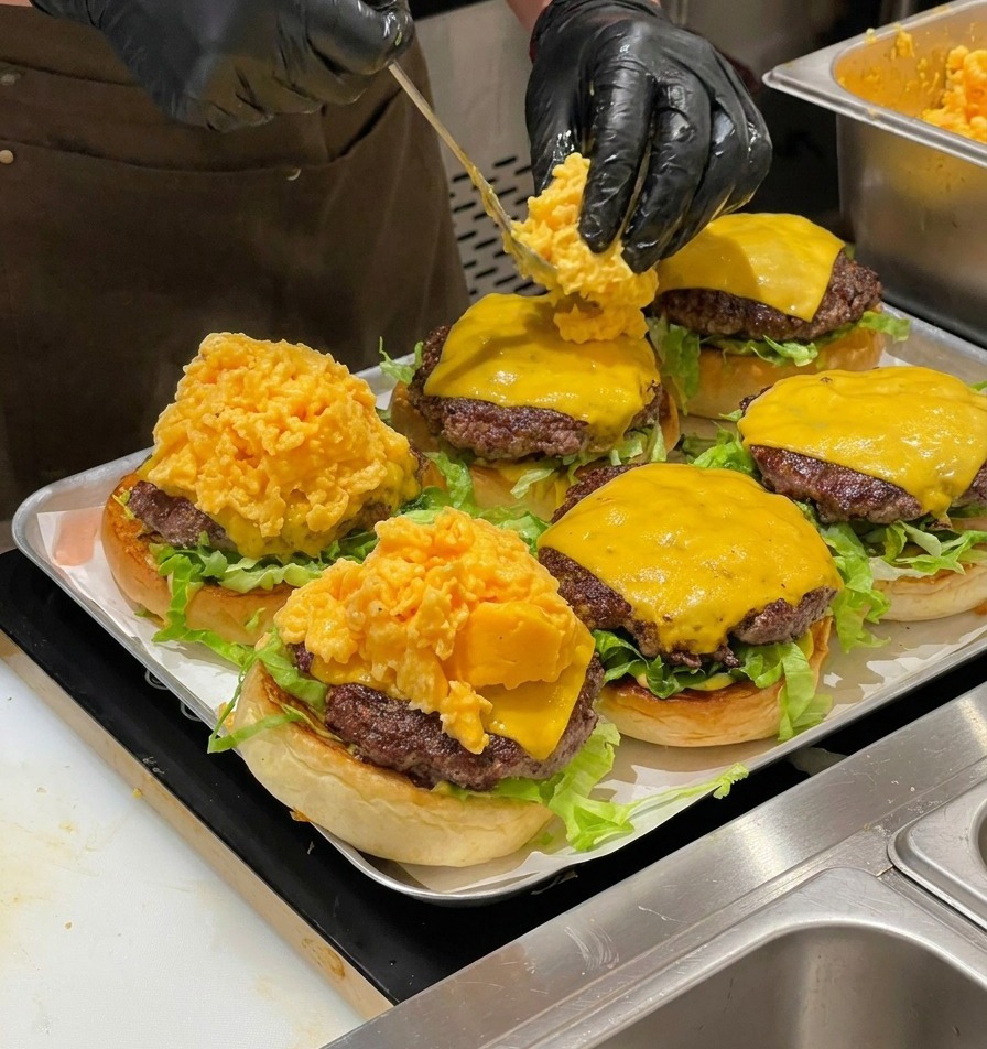 A chef using a spoon to place a large portion of soft scrambled eggs onto cheeseburgers.