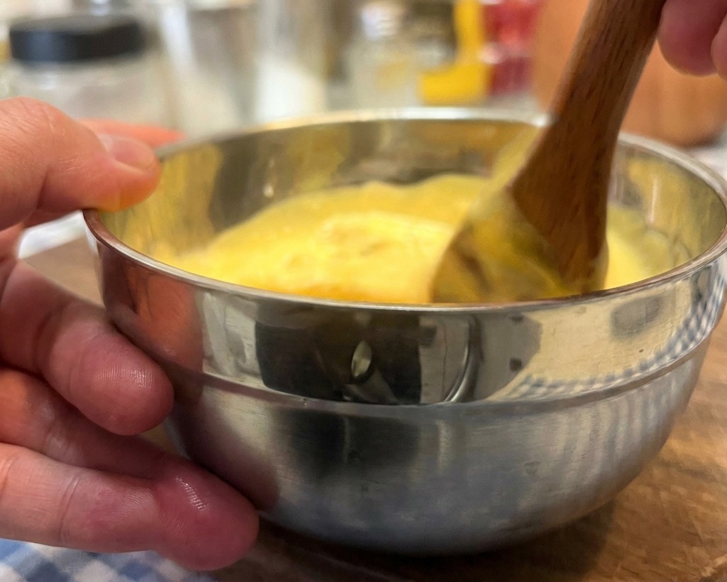 A hand stirring a creamy, yellow-hued burger sauce with a wooden spoon in a small stainless steel bowl.
