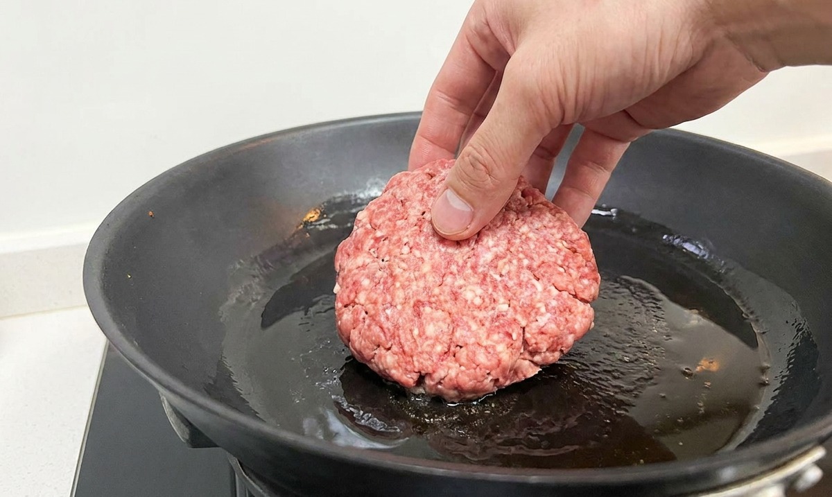 A hand placing a round, raw ground beef patty into a hot black frying pan with shimmering oil.