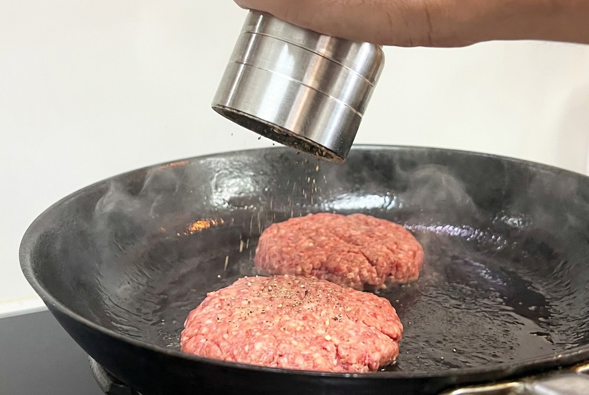 A hand using a metal pepper grinder to season two raw beef patties cooking in a pan.