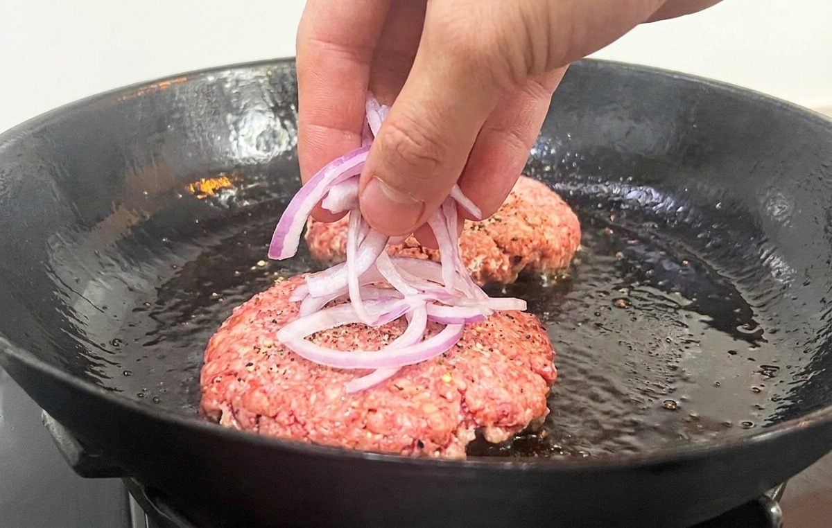 A hand placing thin slivers of red onion on top of raw beef patties in a frying pan.