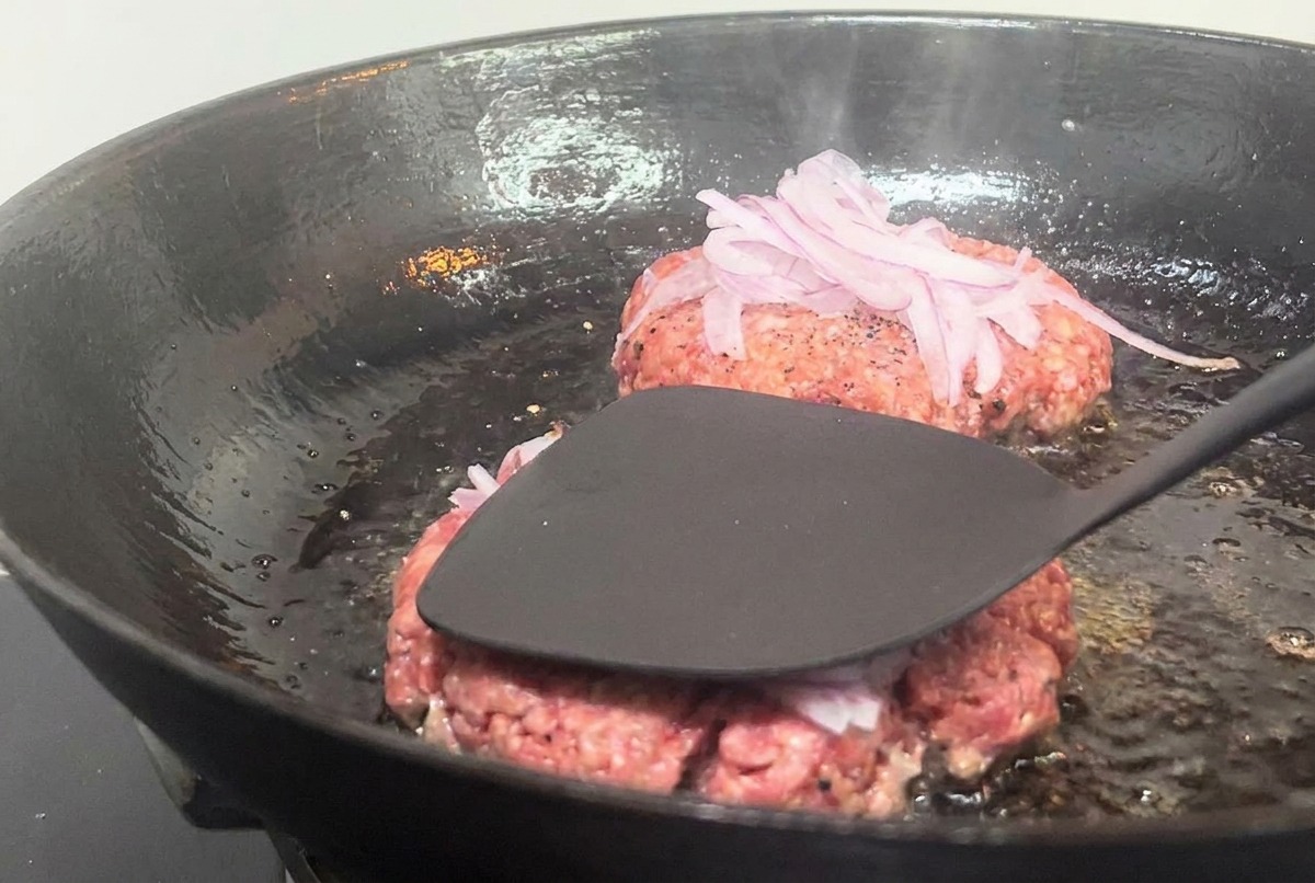 A black metal spatula pressing down on a beef patty topped with onions in a hot pan.