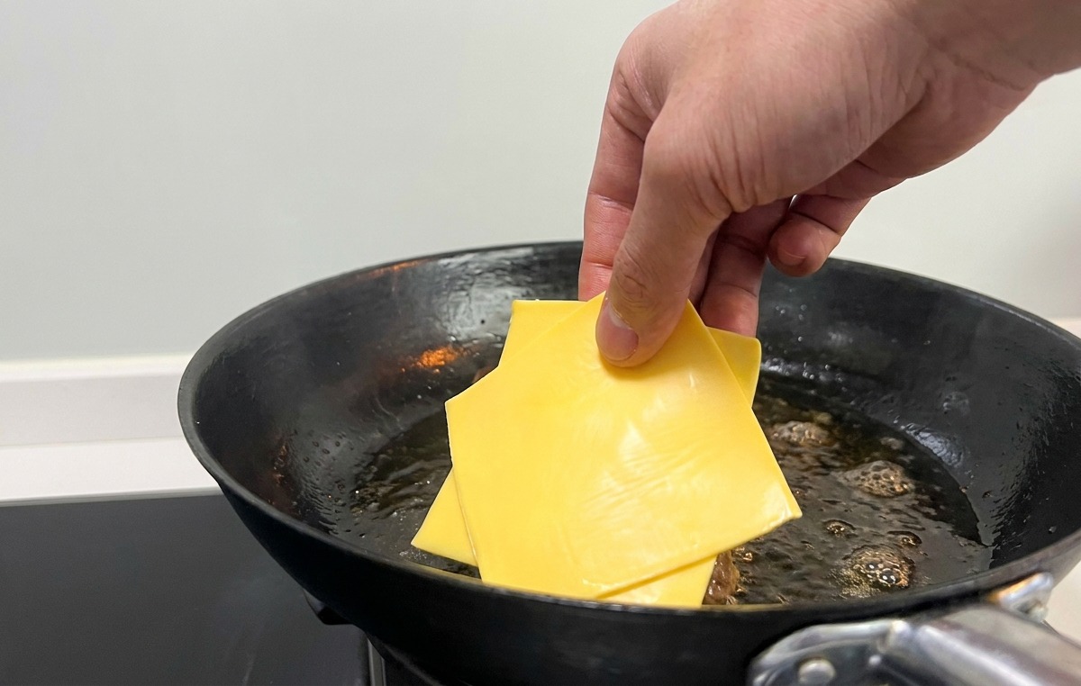 A hand placing square slices of yellow cheese onto seared burger patties in a pan.