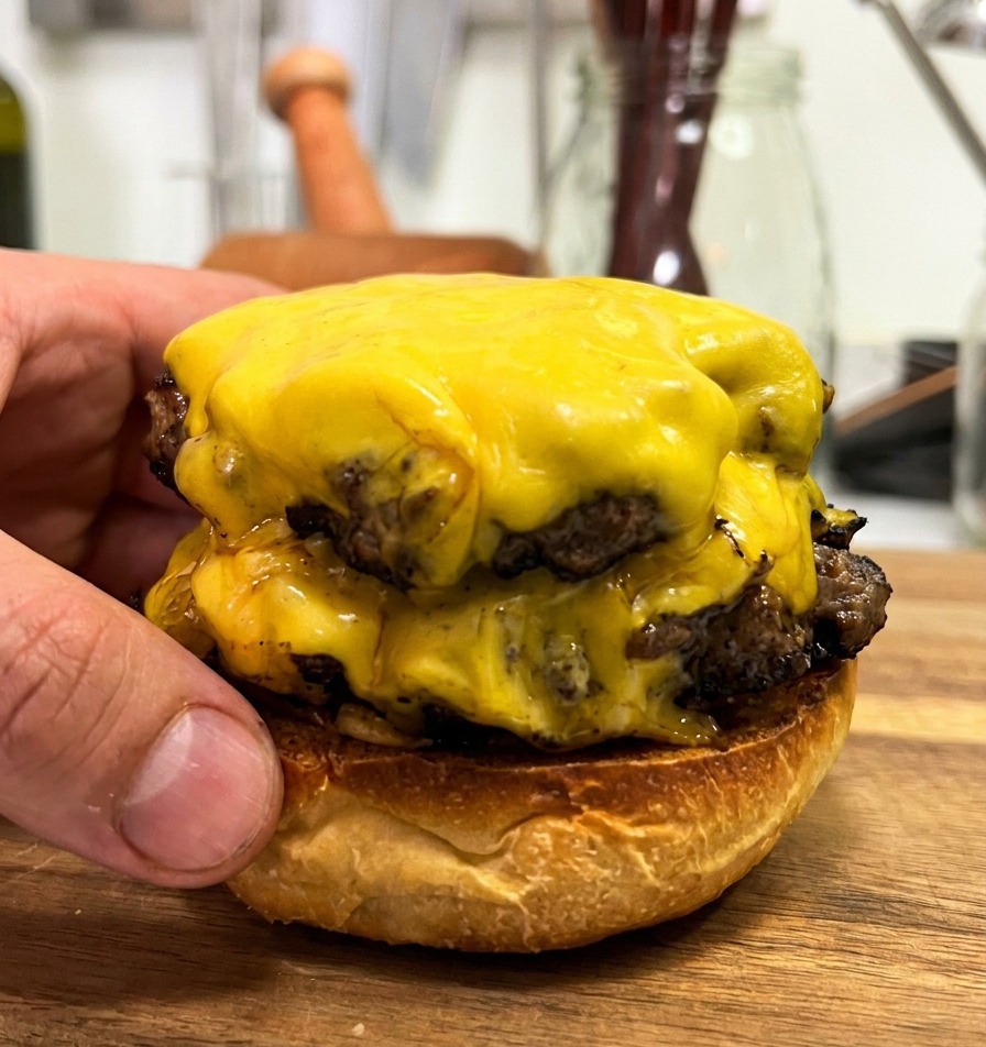 Two thick beef patties with melted yellow cheese being stacked on a toasted bottom bun, showcasing the juicy meat and melting cheese.