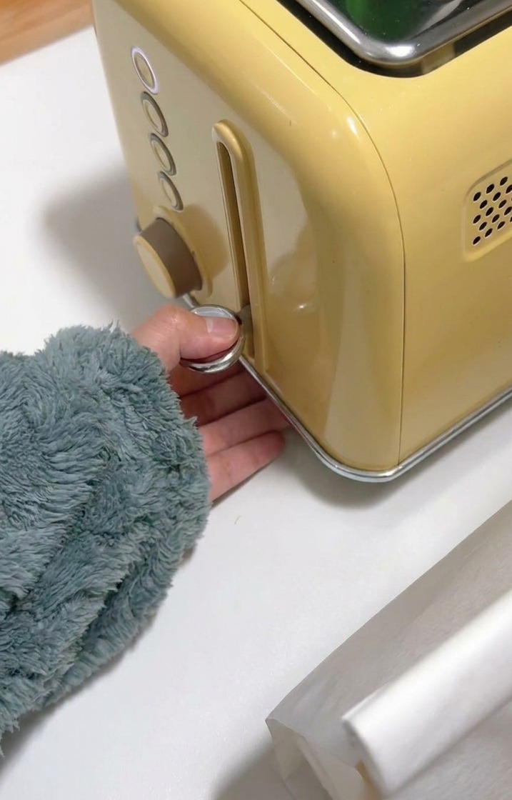 A hand pressing down the chrome lever of a yellow toaster to begin browning the bread.