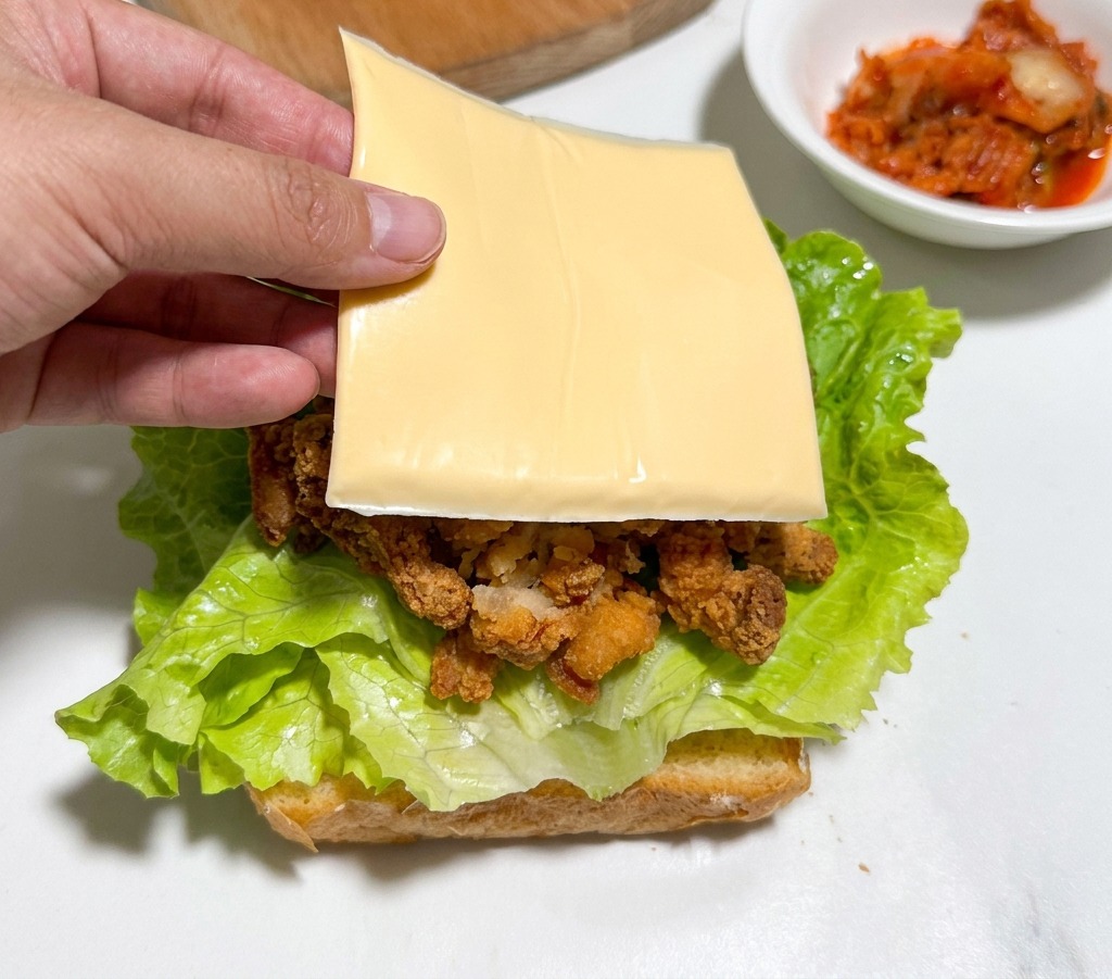 A hand placing a square slice of yellow cheese onto crispy fried chicken pieces layered over green lettuce and a slice of toasted bread.