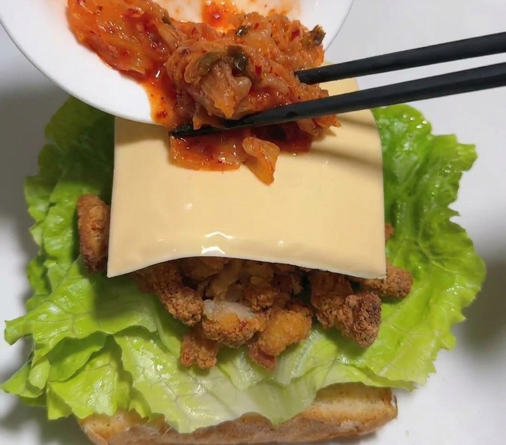 Black chopsticks transferring vibrant red cabbage kimchi from a white bowl onto a slice of yellow cheese layered inside a fried chicken sandwich.