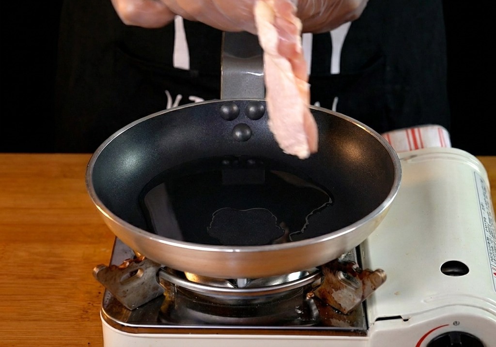 A hand wearing a clear glove placing a raw chicken thigh into a shallow frying pan with heated oil.