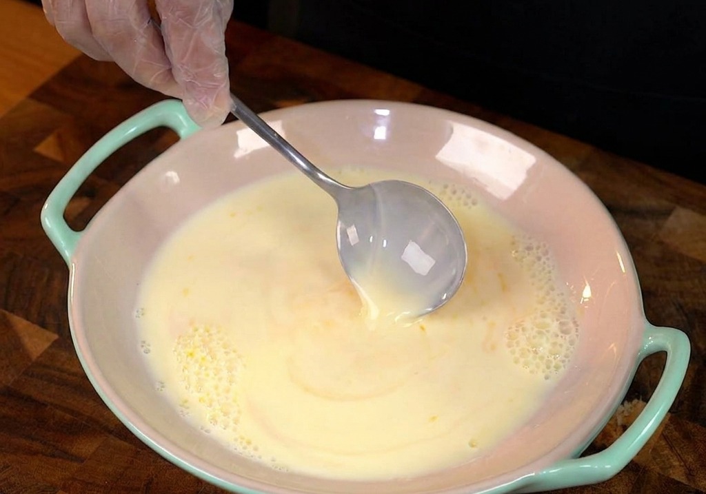 A thick slice of white bread being dipped into a shallow bowl of yellow egg and milk custard.
