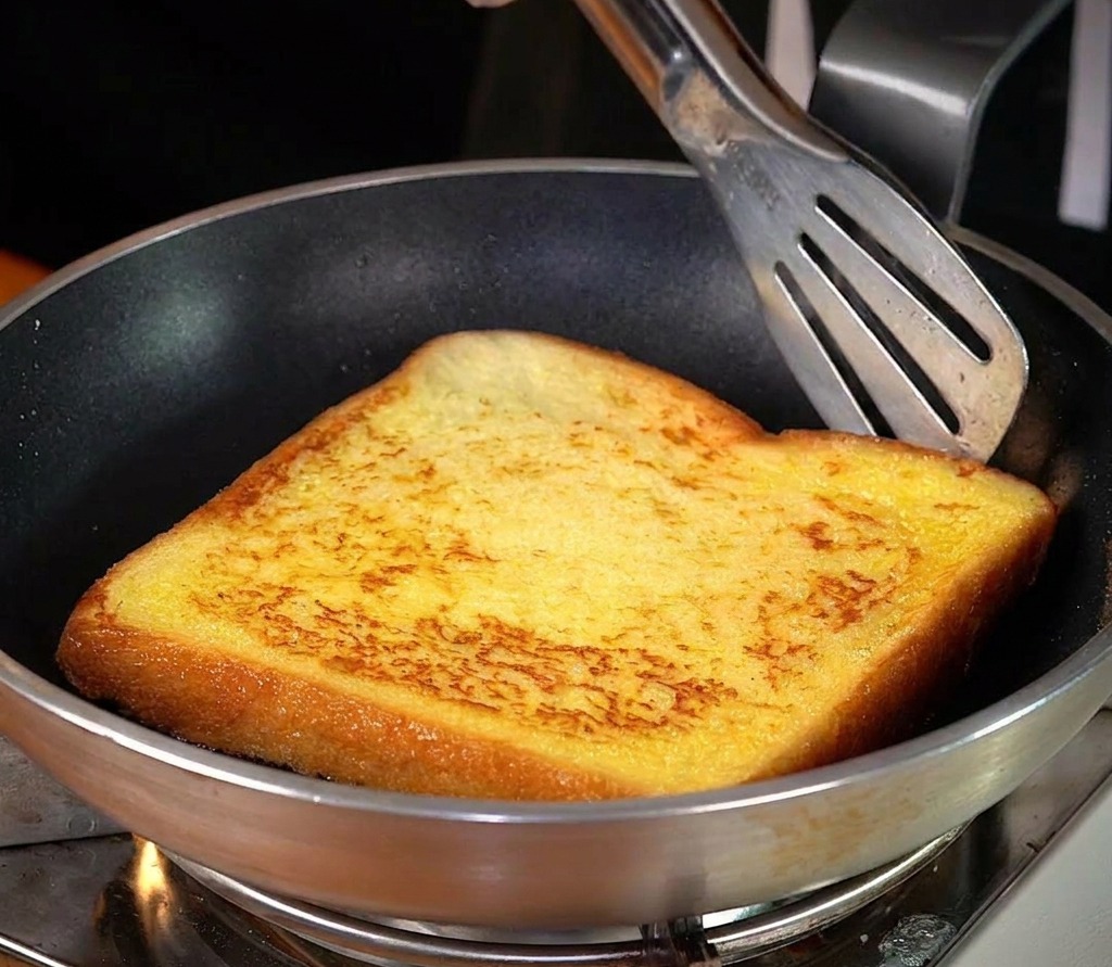 A single slice of thick toast being fried in a silver pan with melted butter, showing a vibrant golden-brown crust.