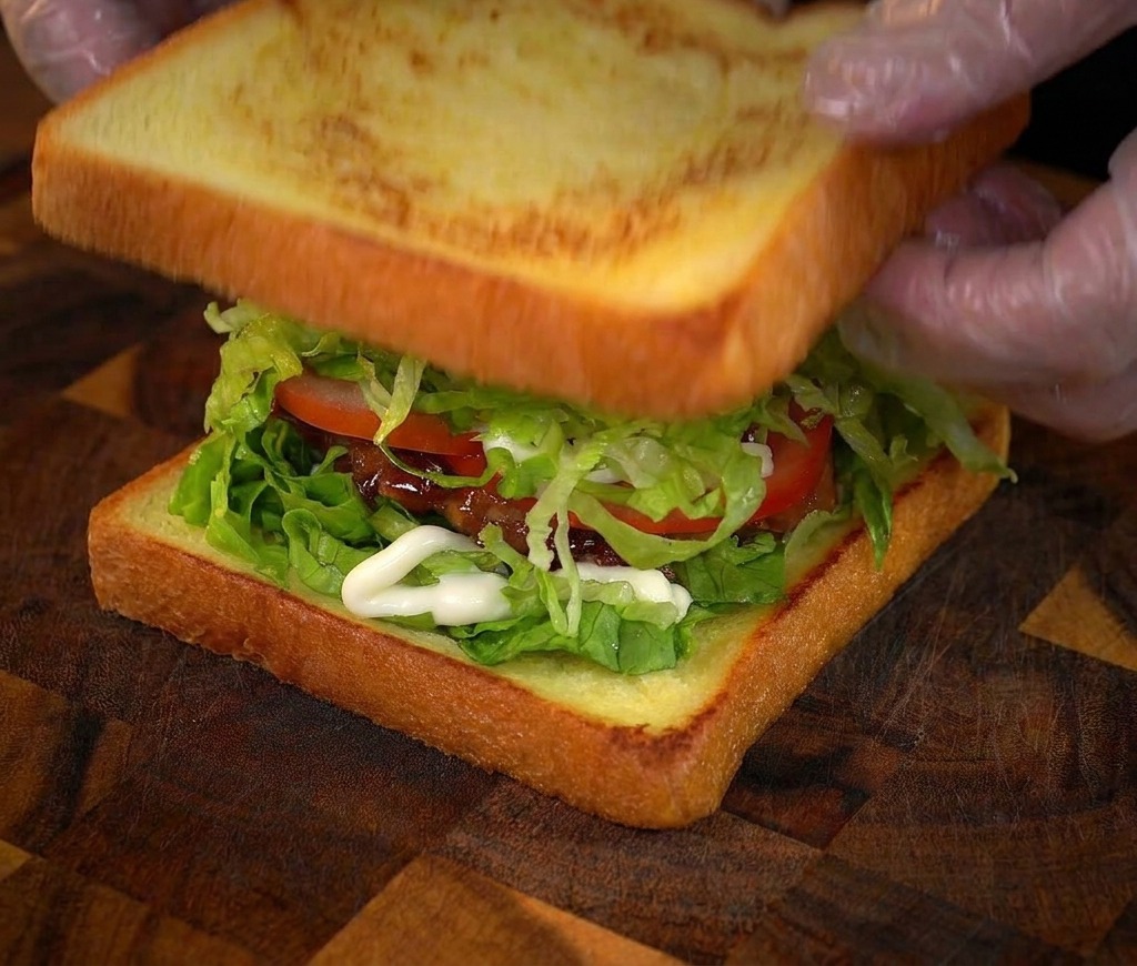 Hands placing the final slice of toasted bread on top of a tall, multi-layered chicken sandwich.