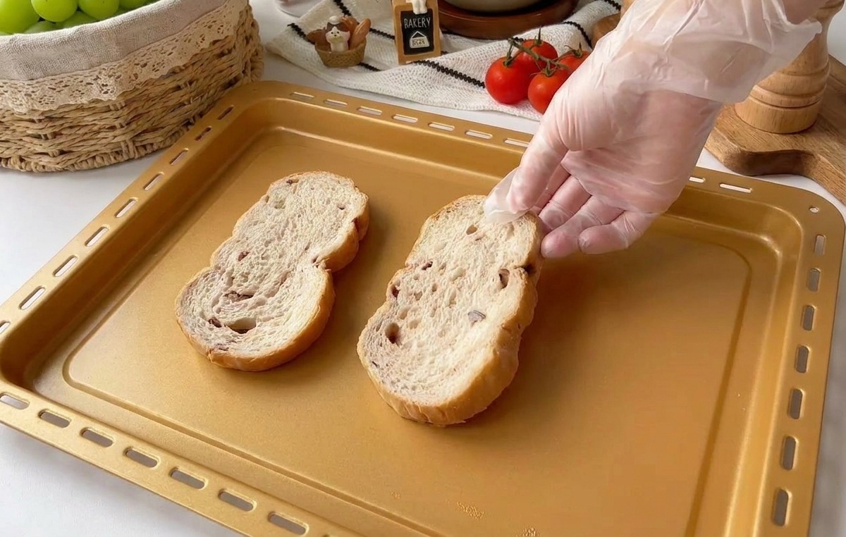 A hand in a clear plastic glove placing two thick slices of rustic multigrain bread onto a gold-colored rectangular baking sheet.