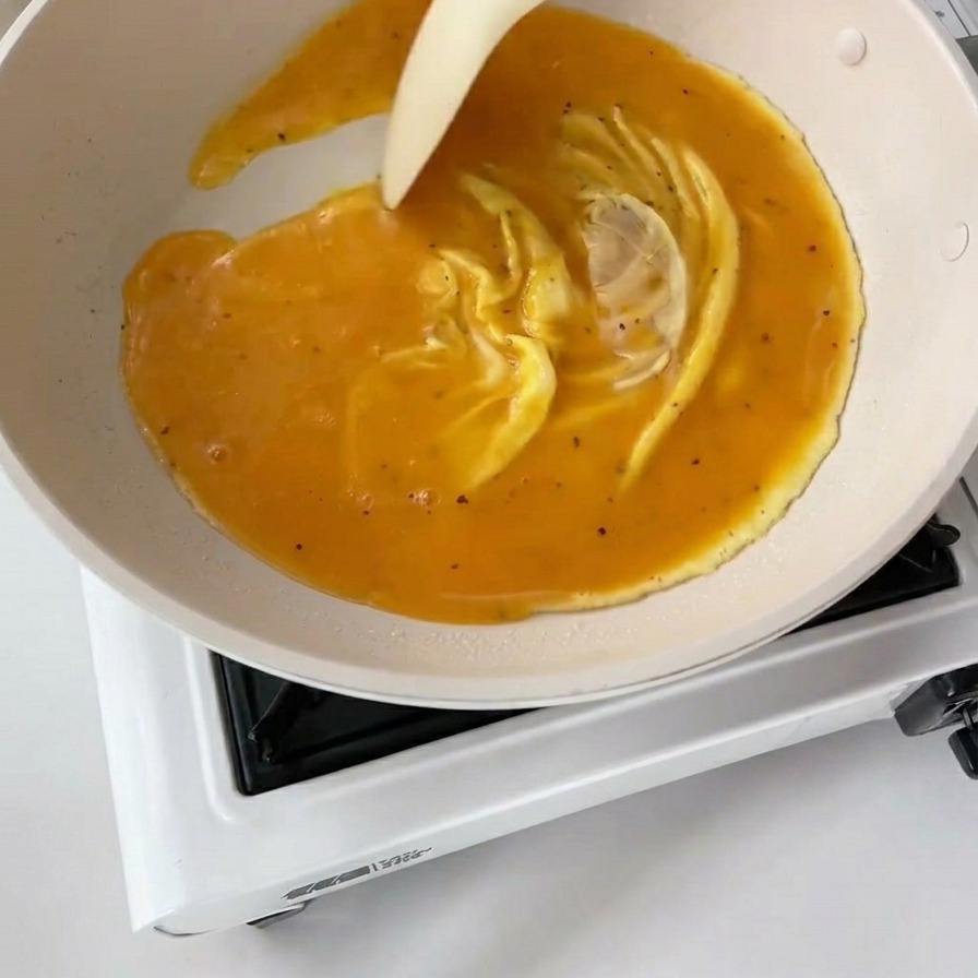 A white spatula pushing through partially set eggs in a white pan, creating thick, wavy yellow folds.