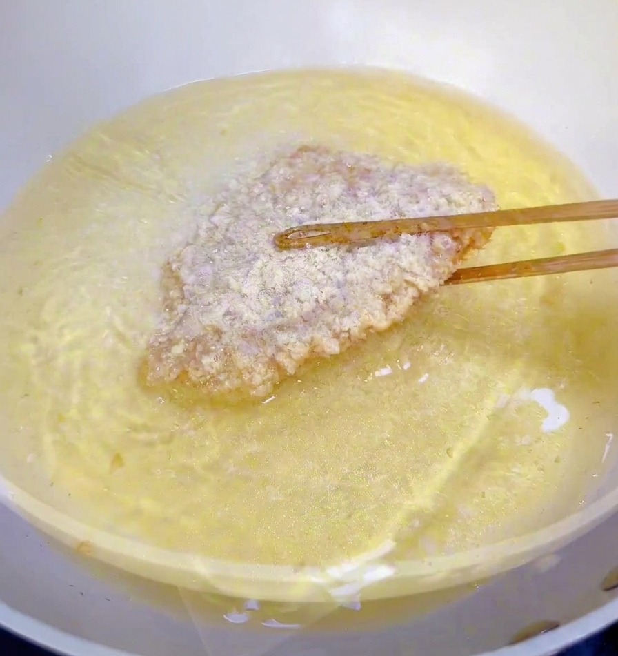 A piece of breaded chicken fillet being lowered into a pan of shimmering hot oil with wooden chopsticks.