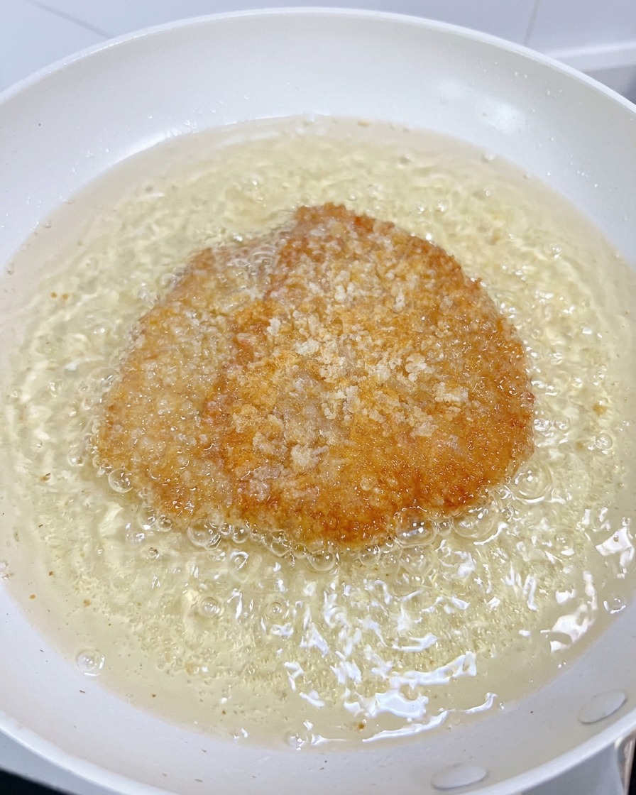 A chicken fillet deep-frying in a white pan of bubbling oil, its coating turning a crispy golden brown.