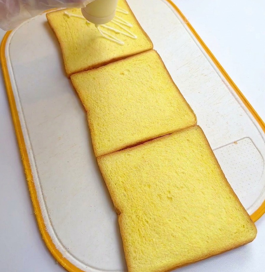 A person wearing plastic gloves drizzling white salad dressing from a squeeze bottle over three square slices of yellow toast.