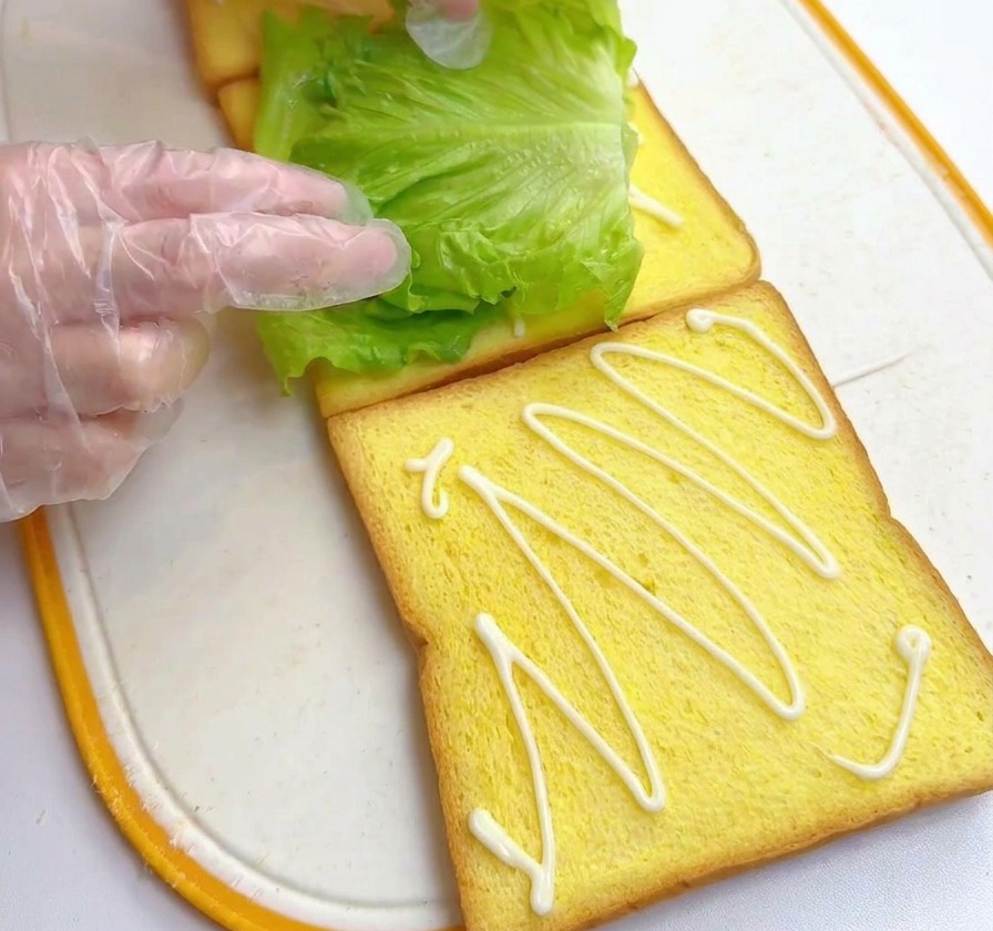Hands placing a vibrant green lettuce leaf onto a slice of yellow toast that has been drizzled with dressing.