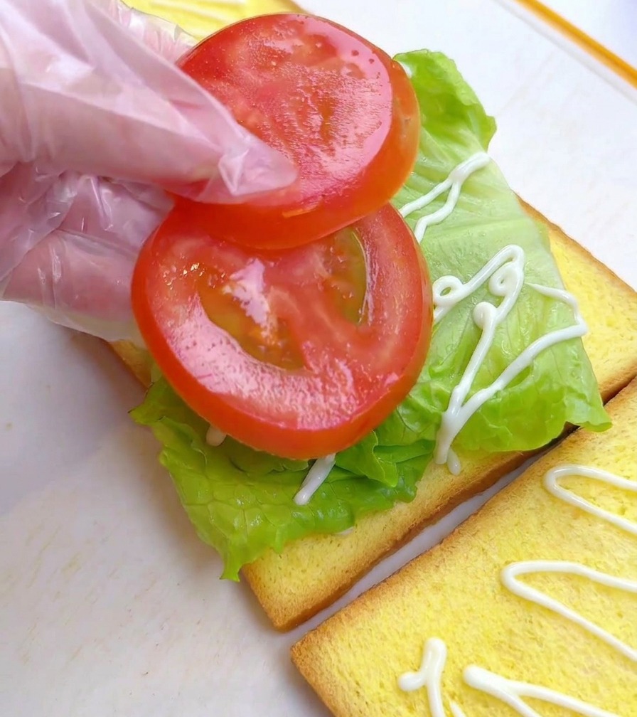 A person wearing a plastic glove placing two round tomato slices onto a bed of green lettuce on a slice of toast.