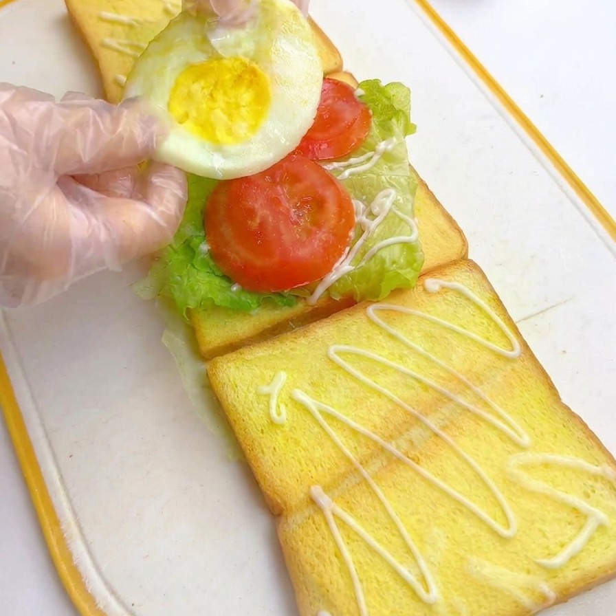 Gloved hands placing a round fried egg onto sliced tomatoes in a multi-layered sandwich assembly.