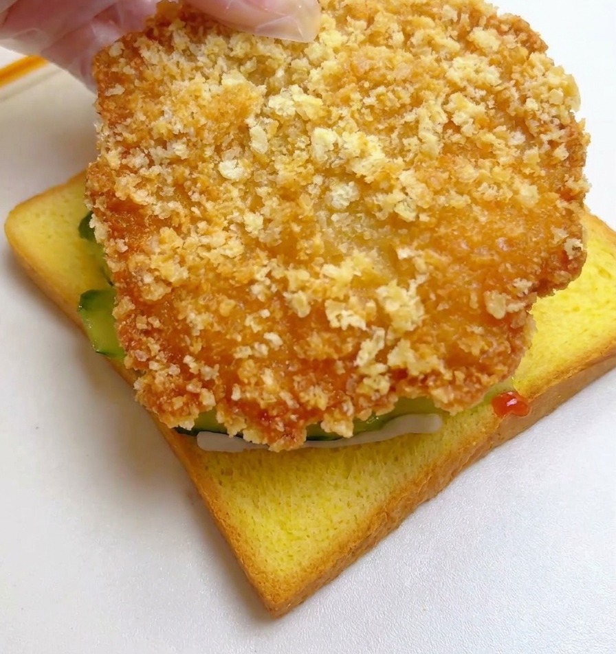 A hand placing a large, breaded and fried golden-brown chicken fillet onto the cucumber-topped toast slice.