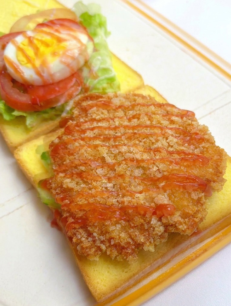 A breaded, golden-brown chicken fillet on toast being drizzled with red ketchup, with other sandwich layers visible in the background.