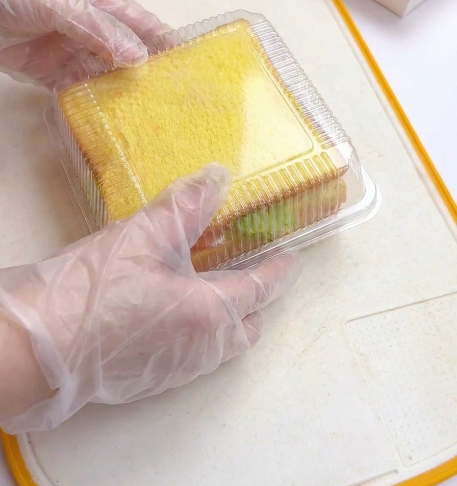 Hands in plastic gloves pressing a clear plastic square lid onto a thick stacked sandwich inside a mold.
