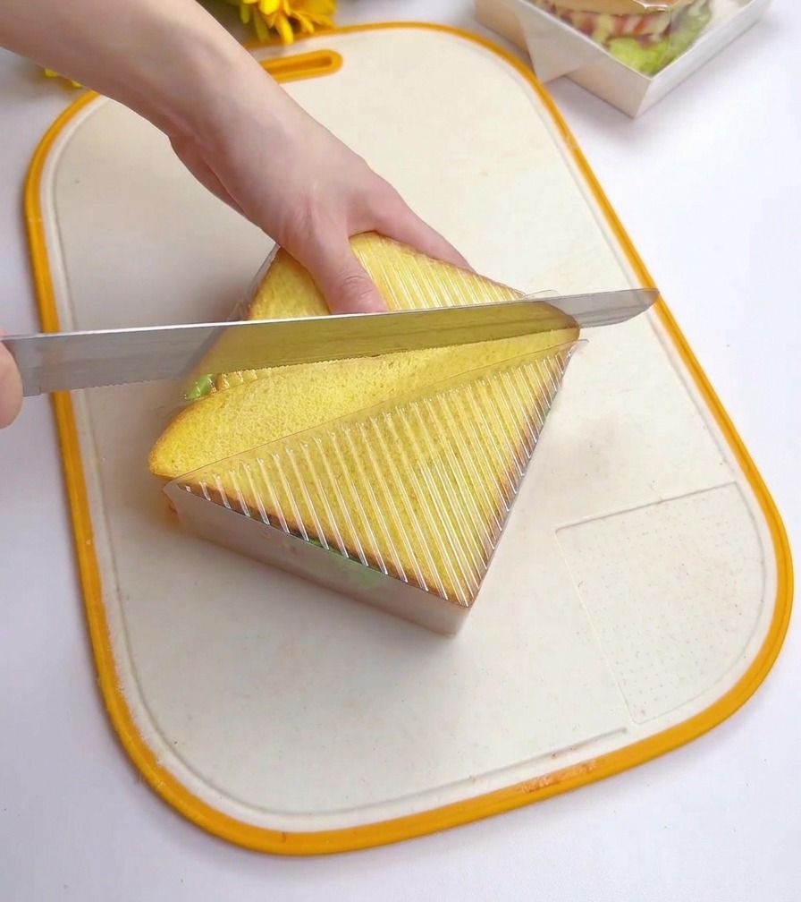 A person using a serrated bread knife to cut a stacked sandwich diagonally while it is held inside a clear plastic triangular container.