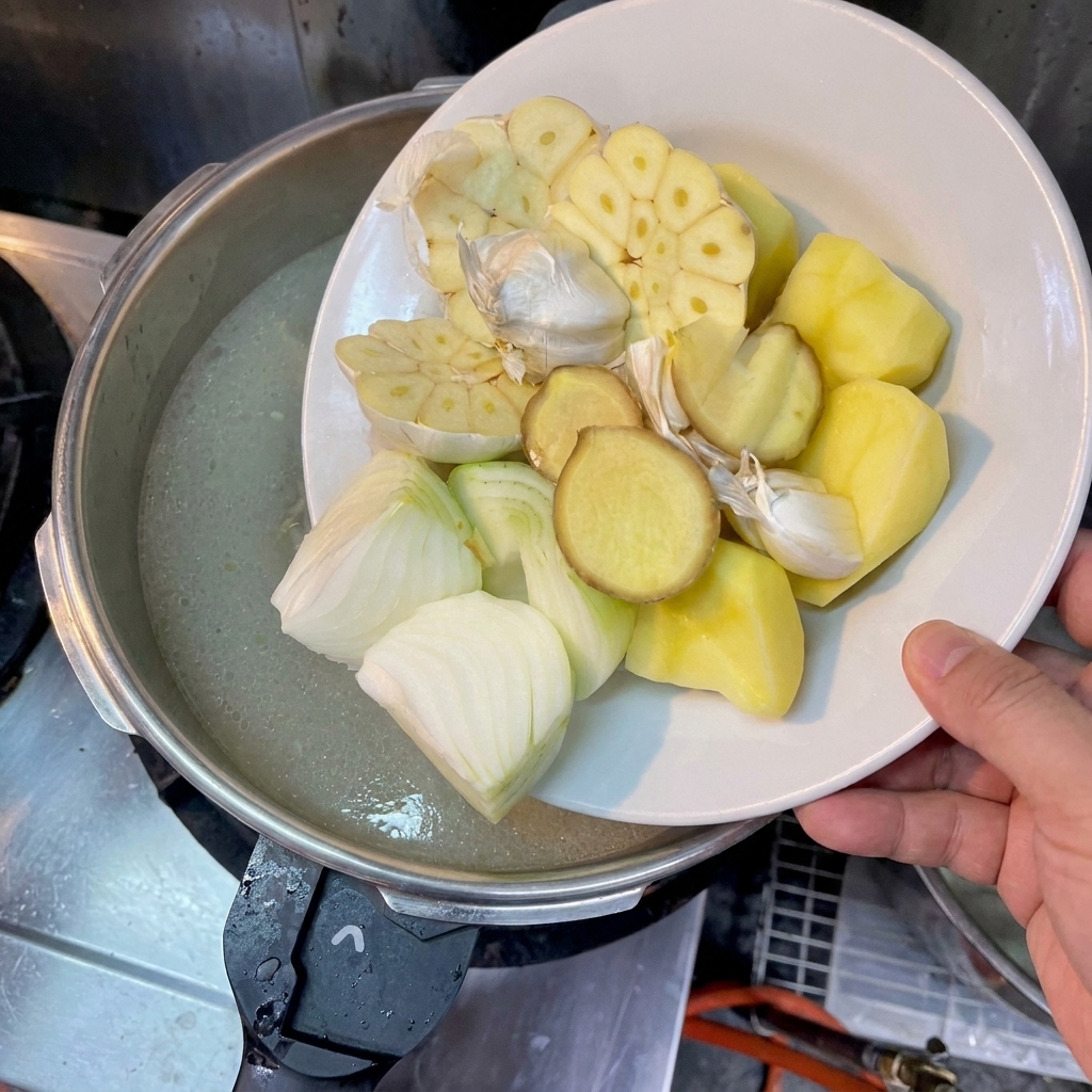 A persons hand holding a white plate, sliding large pieces of potato, halved onions, sliced ginger, and halved garlic heads into a pot of cloudy broth.