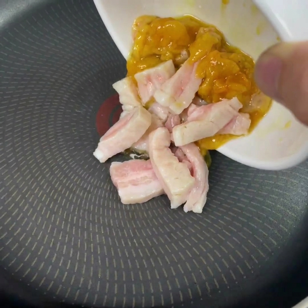 A hand pouring raw pieces of white and yellow fat from a small white bowl into a cold, dry frying pan.
