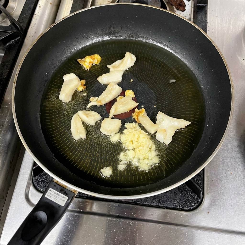 Minced garlic and pieces of rendering fat cooking in a shallow pool of hot, clear oil in a frying pan.