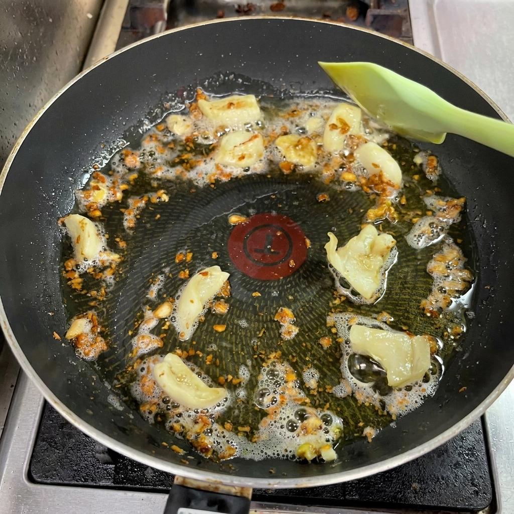 Minced garlic frying in oil with pieces of fat in a non-stick pan, turning a deep golden brown, while being gently stirred by a green spatula.