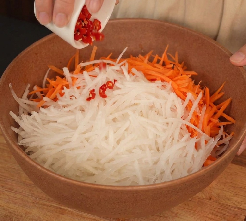 A bowl containing shredded white radish and orange carrots with chopped red chilies being added from a small white dish.