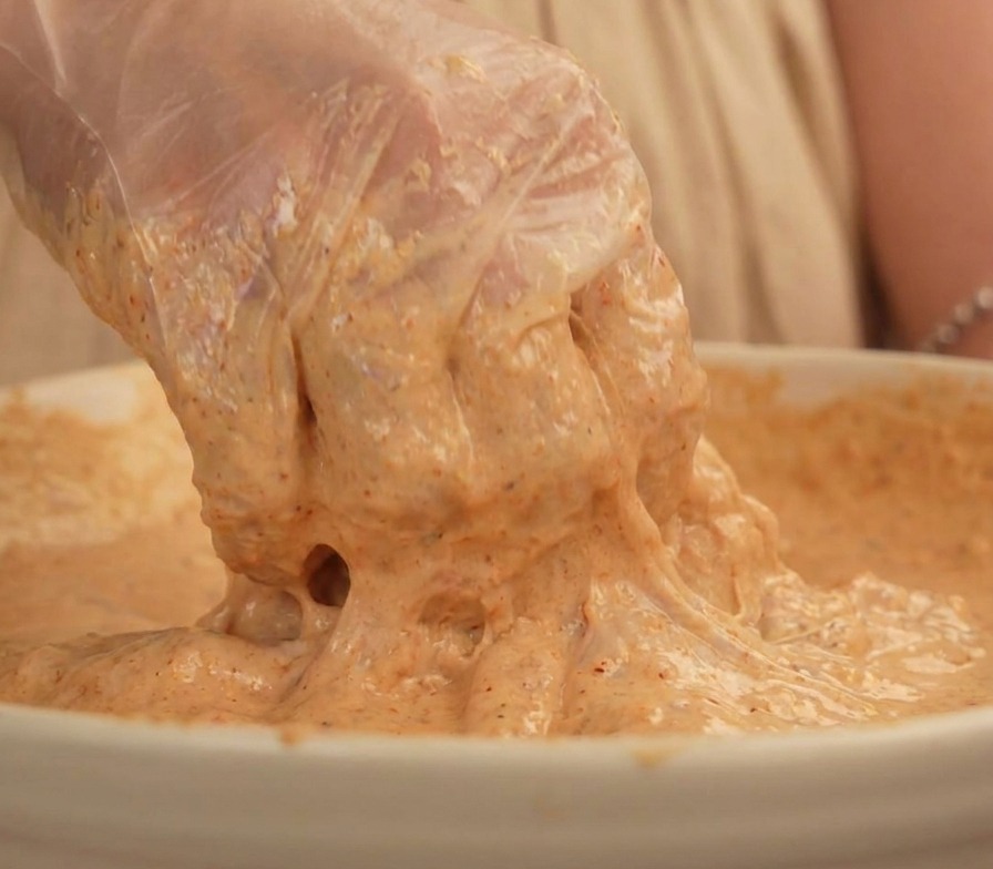 A hand in a clear plastic glove lifting a piece of chicken thigh completely coated in a thick beige colored batter.